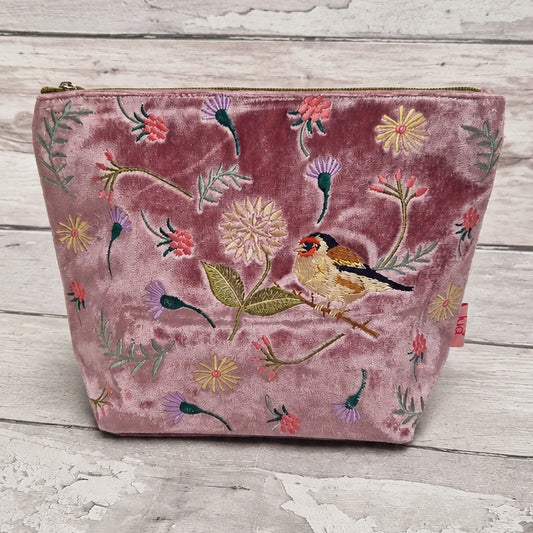 A dusty rose velvet zippered pouch decorated with colorful embroidered flowers, leaves, and a small goldfinch bird, displayed on a rustic white-washed wooden surface.