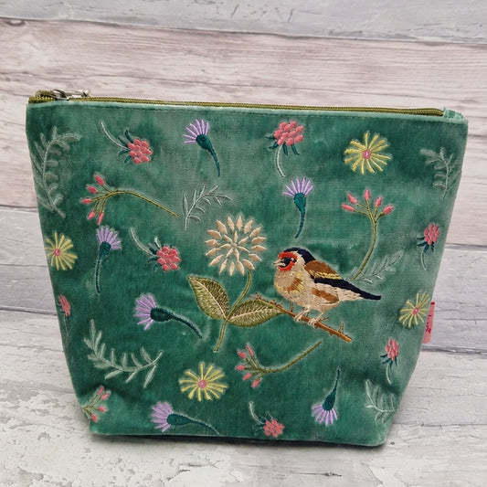 A green velvet zippered pouch decorated with colorful embroidered flowers and a goldfinch bird, set against a light gray wooden background.