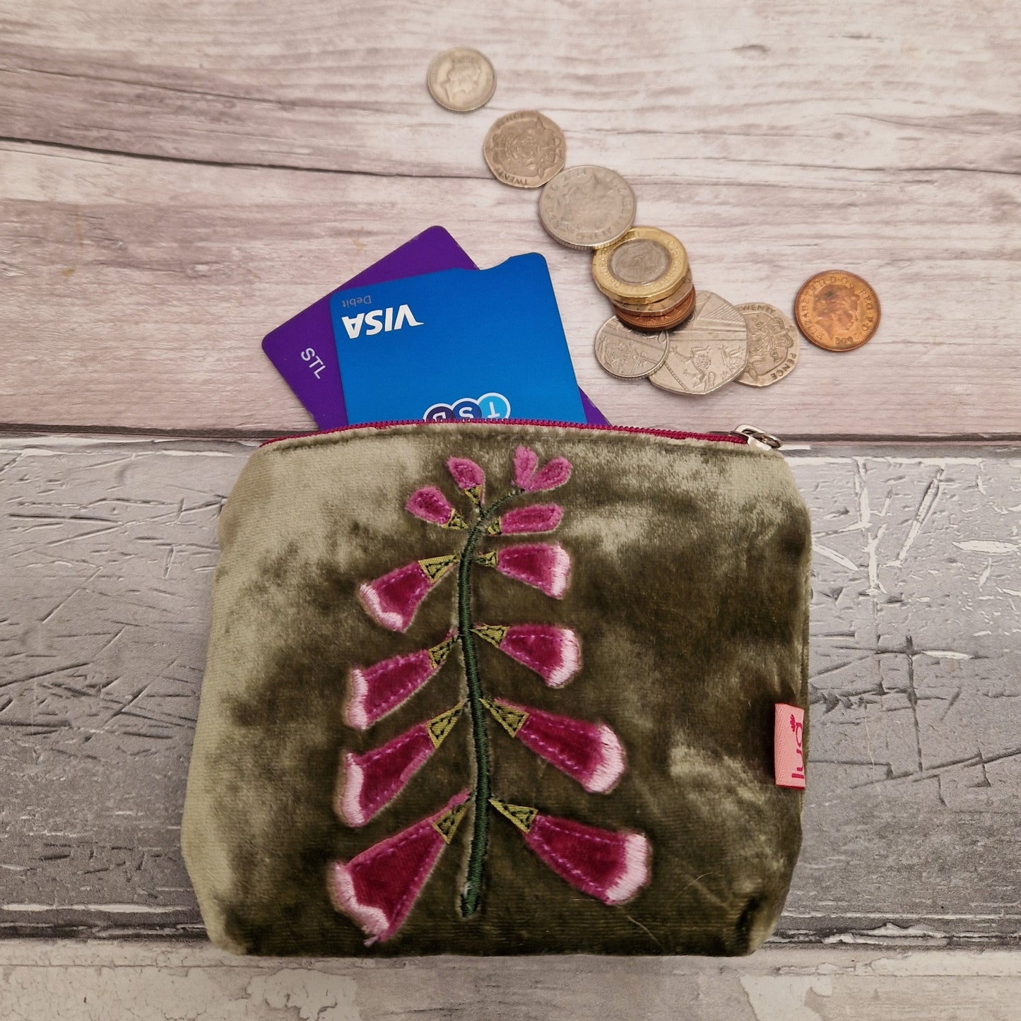 Green velvet coin purse decorated with a pink foxglove stem, filled with coins and cards.