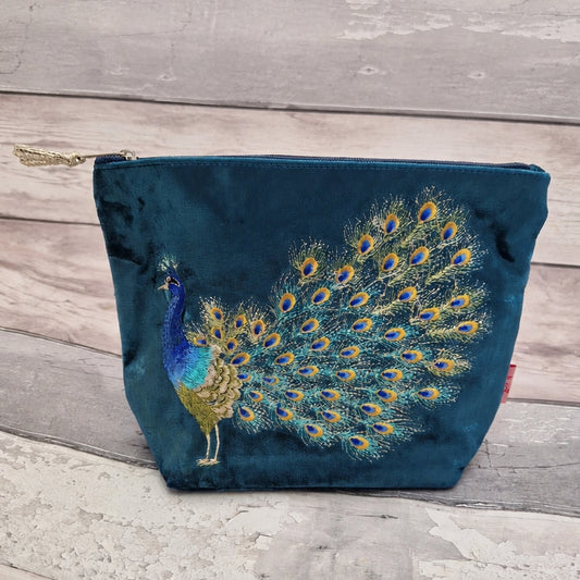 A teal velvet cosmetic pouch featuring an embroidered peacock with a fully fanned tail of blue, gold, and green feathers, resting on a light gray wooden plank surface.