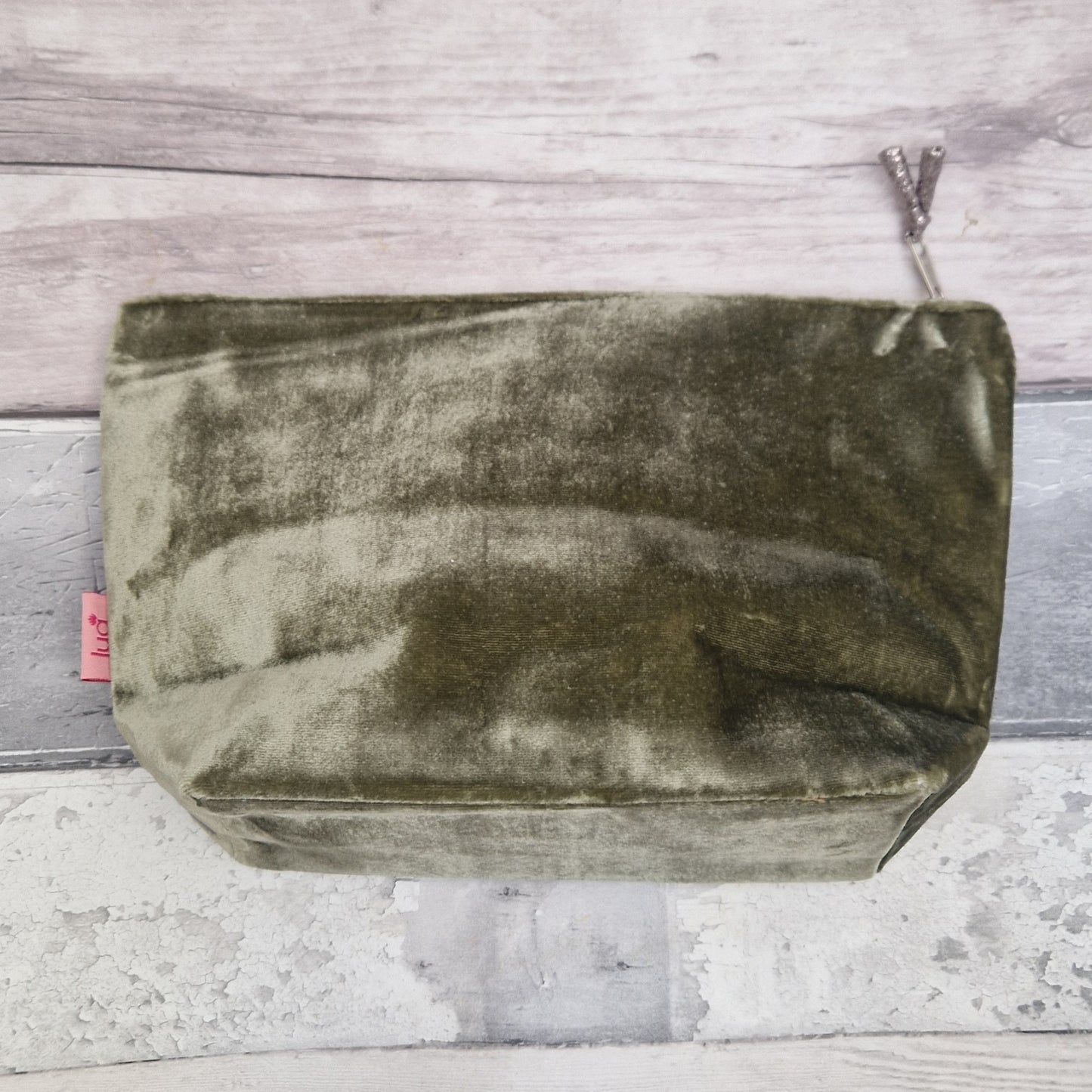 An olive green velvet zippered pouch with a silver zipper pull, photographed from above on a rustic gray and white wood plank background.