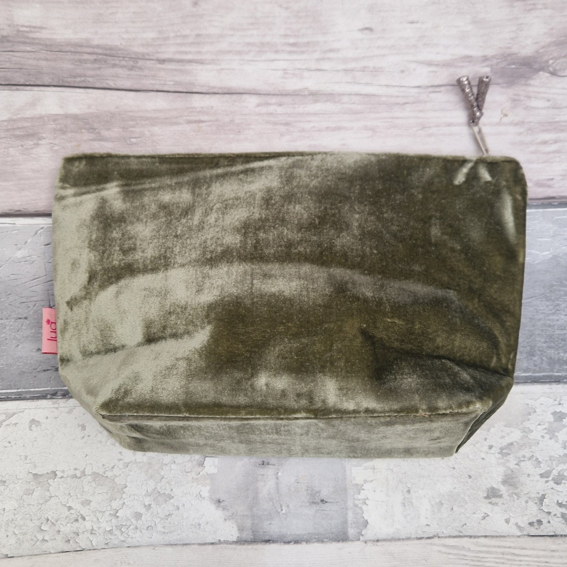 An olive green velvet zippered pouch with a silver zipper pull, photographed from above on a rustic gray and white wood plank background.