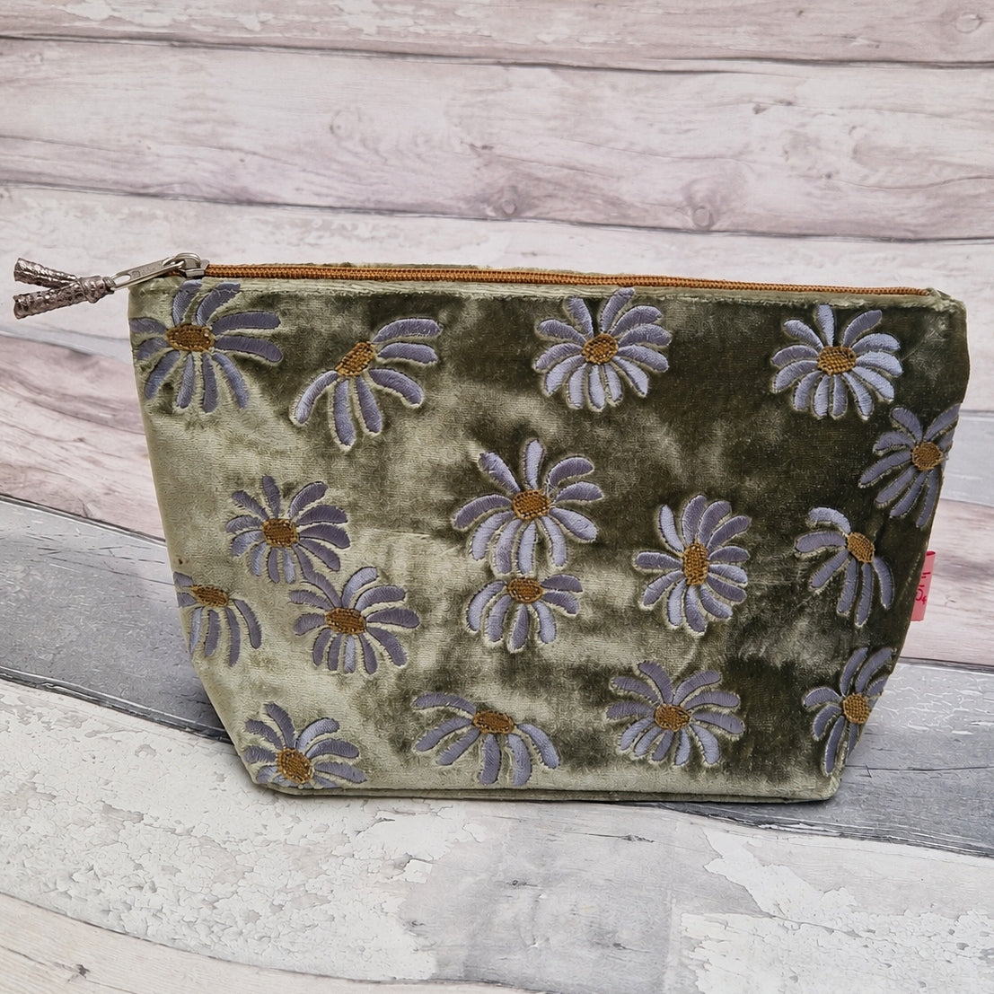 An olive green velvet zipper pouch decorated with a pattern of lavender-gray and gold embroidered daisies, set against a rustic whitewashed wood background.