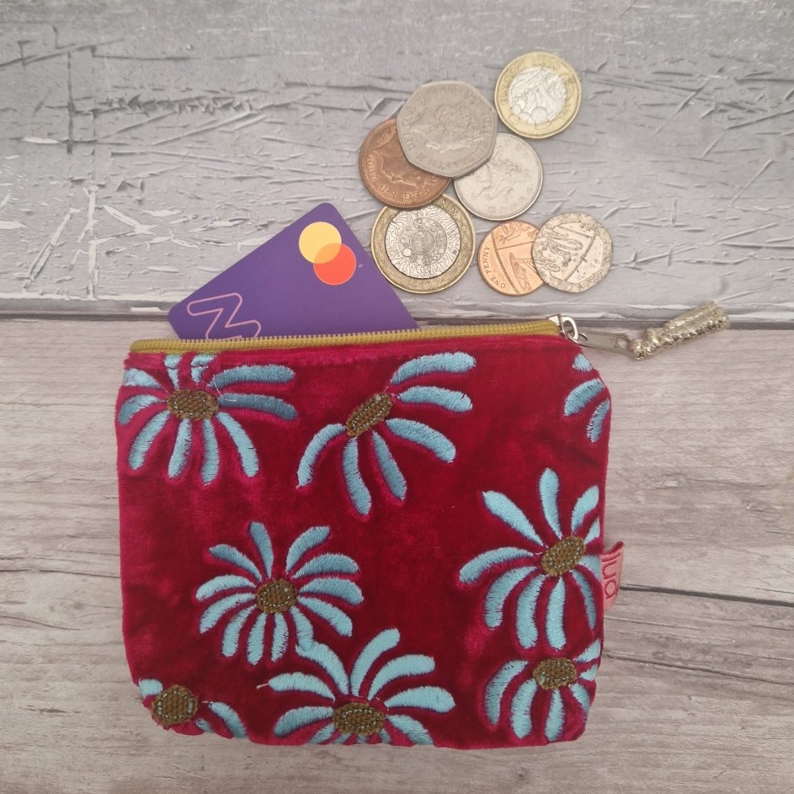 A hot pink velvet zippered pouch with embroidered daisy flowers, shown on a gray wood-textured background with coins and a card peeking out from the top.