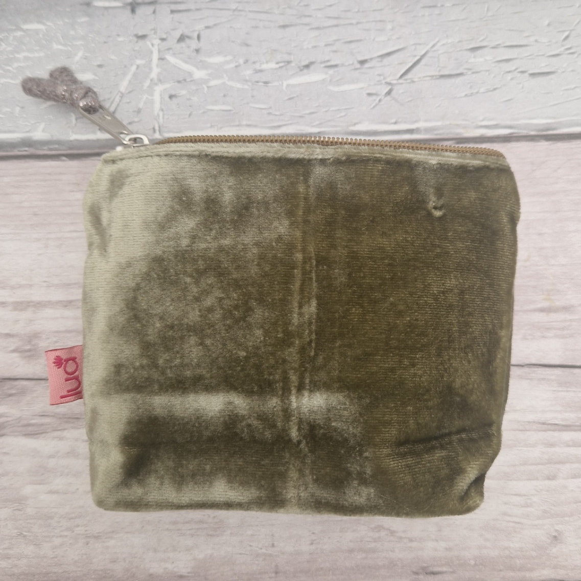 An olive green velvet zippered pouch with a small pink tag on the side, resting on a light gray wooden surface.