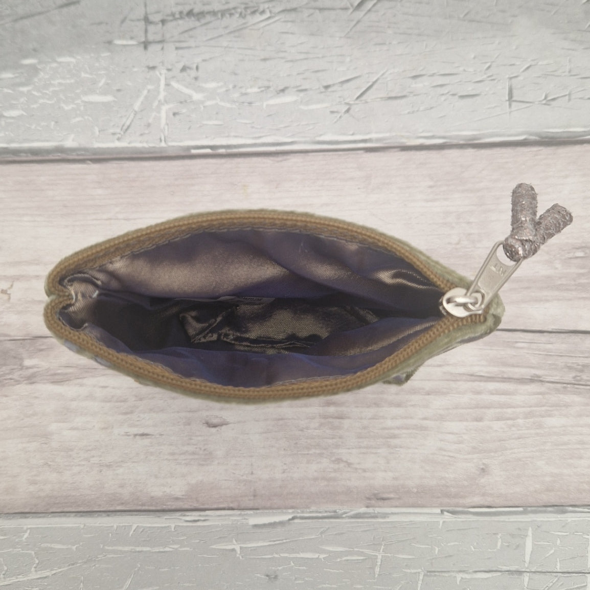 An olive green zippered pouch with a silk lining, shown open on a light gray wood plank background.