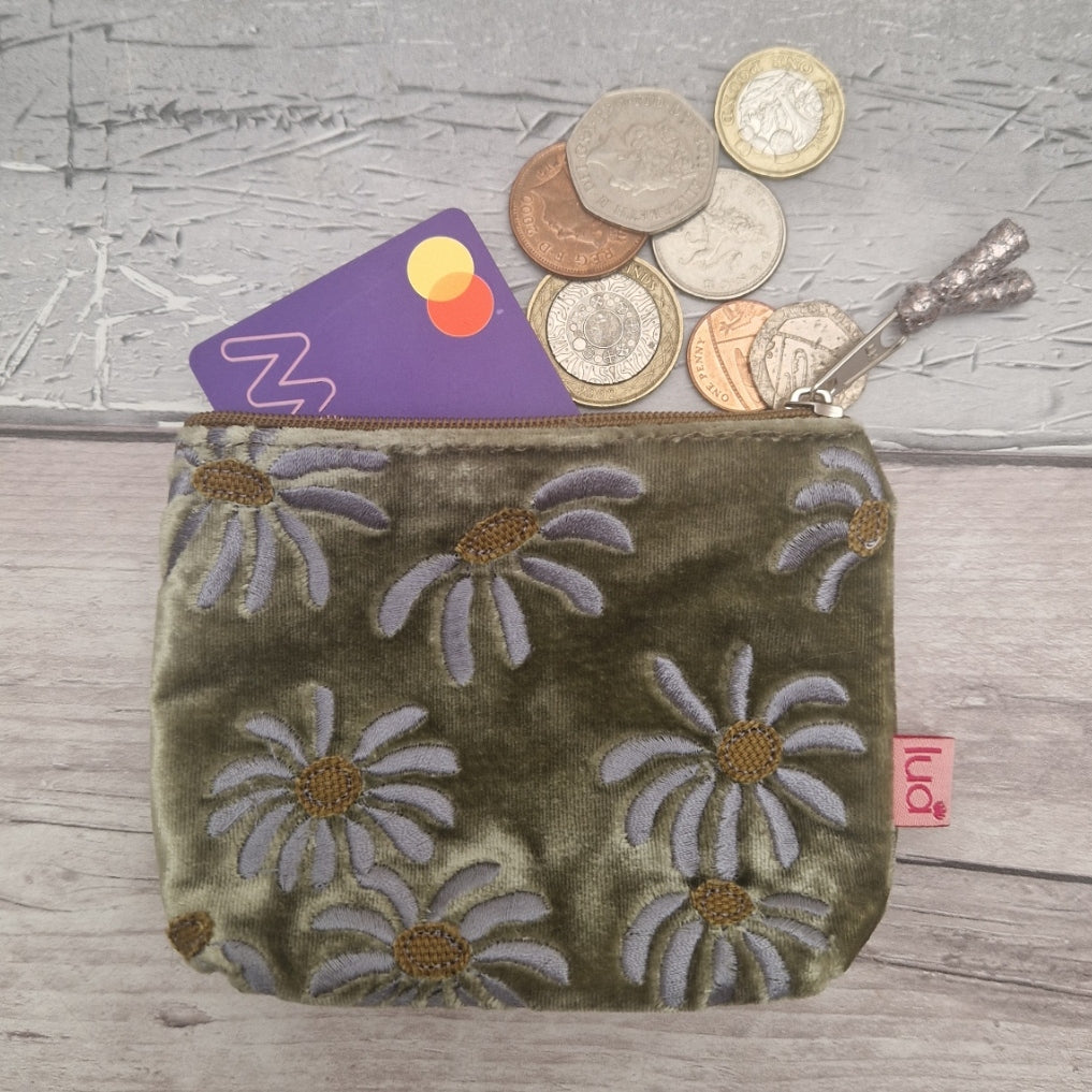 An olive green velvet coin purse with a daisy pattern, shown with a card and coins protruding from the top zipper. A small pink tag on the right side reads 'lua'.