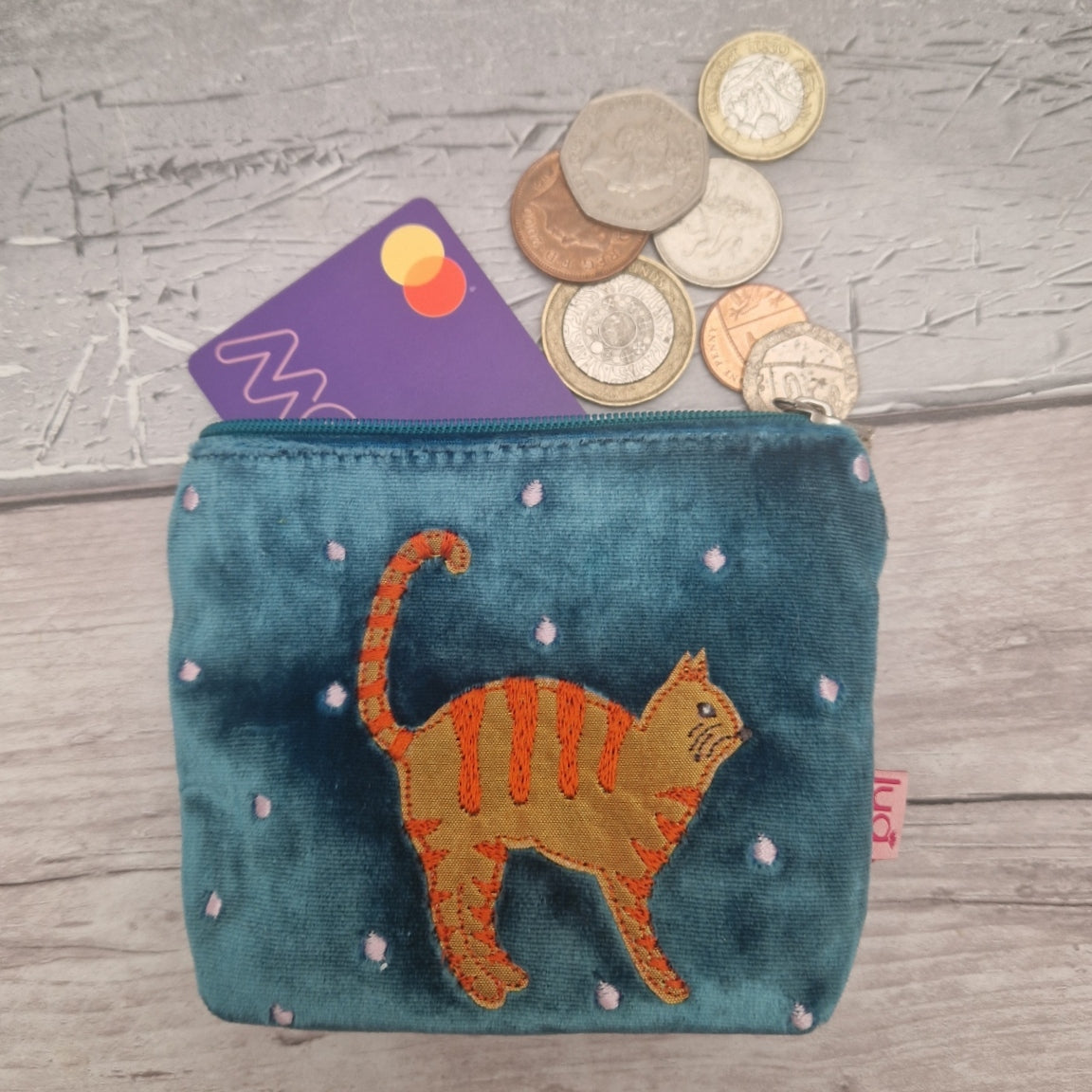 A small turquoise velvet coin purse with an orange embroidered cat design on the front, shown open with coins and a card sticking out. The purse is placed on a light gray wood-textured surface.