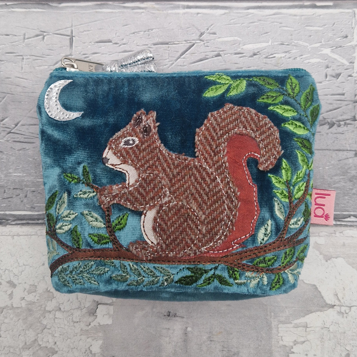 A teal velvet zippered pouch featuring an embroidered brown and red squirrel on a tree branch with green leaves and a silver crescent moon, shown against a light gray textured background.