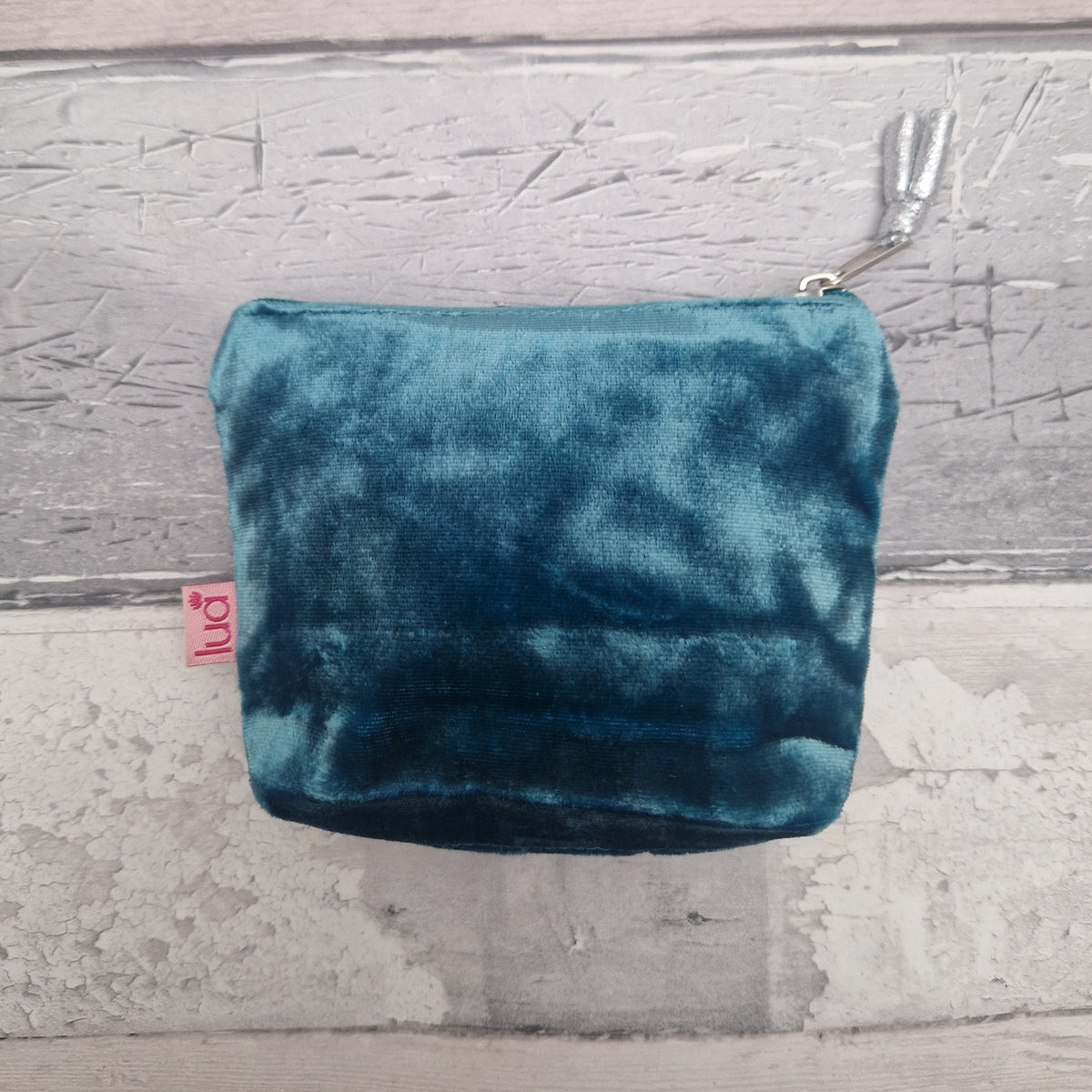 A teal blue velvet zippered pouch with a small pink 'lua' brand tag, resting on a rustic, light gray wooden plank surface.
