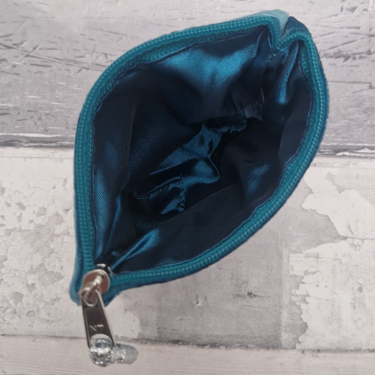 A small teal zippered pouch with a shiny teal satin interior and a silver metal zipper pull, shown open against a distressed light gray wood background.