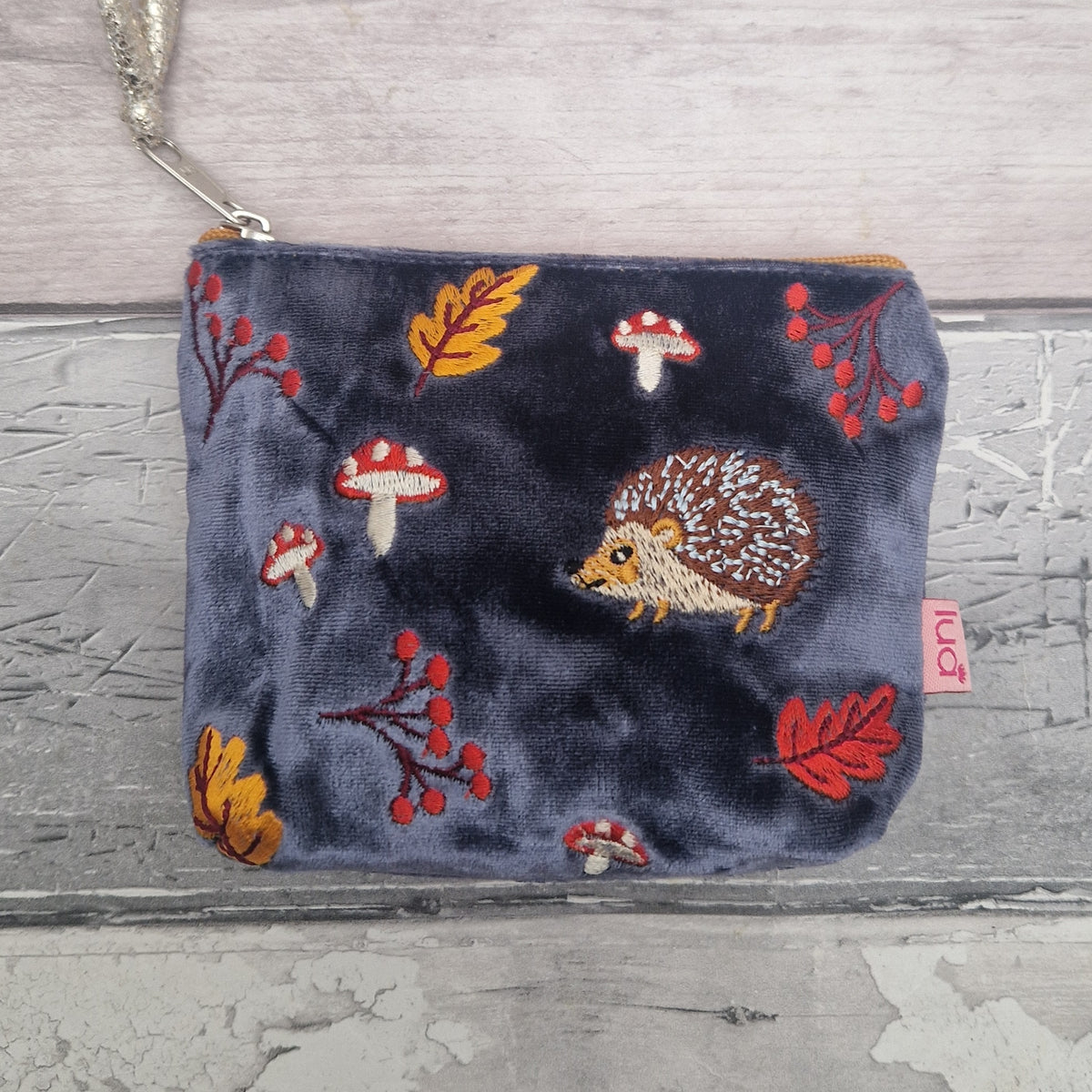 A small zip pouch in dark navy blue velvet with embroidered woodland motifs including a hedgehog, mushrooms, leaves, and berries, shown on a rustic gray wood background. A small pink tag on the right side reads 'Lua'.