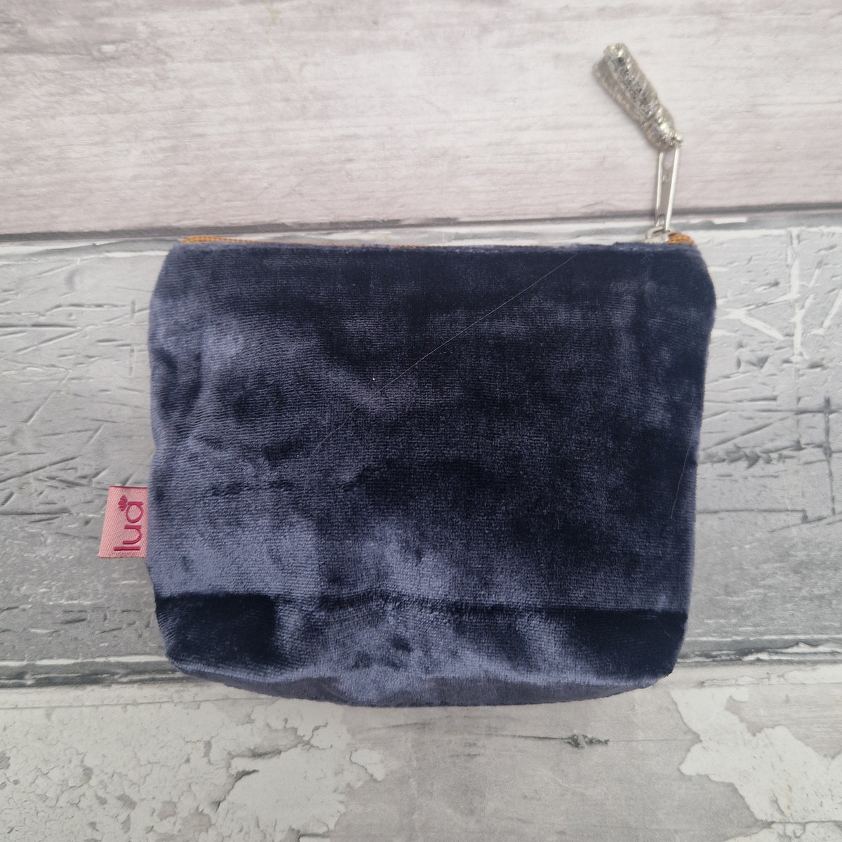 A small navy blue velvet zippered pouch with a silver zipper pull and a small pink 'lua' brand tag, displayed on a rustic gray wooden background.
