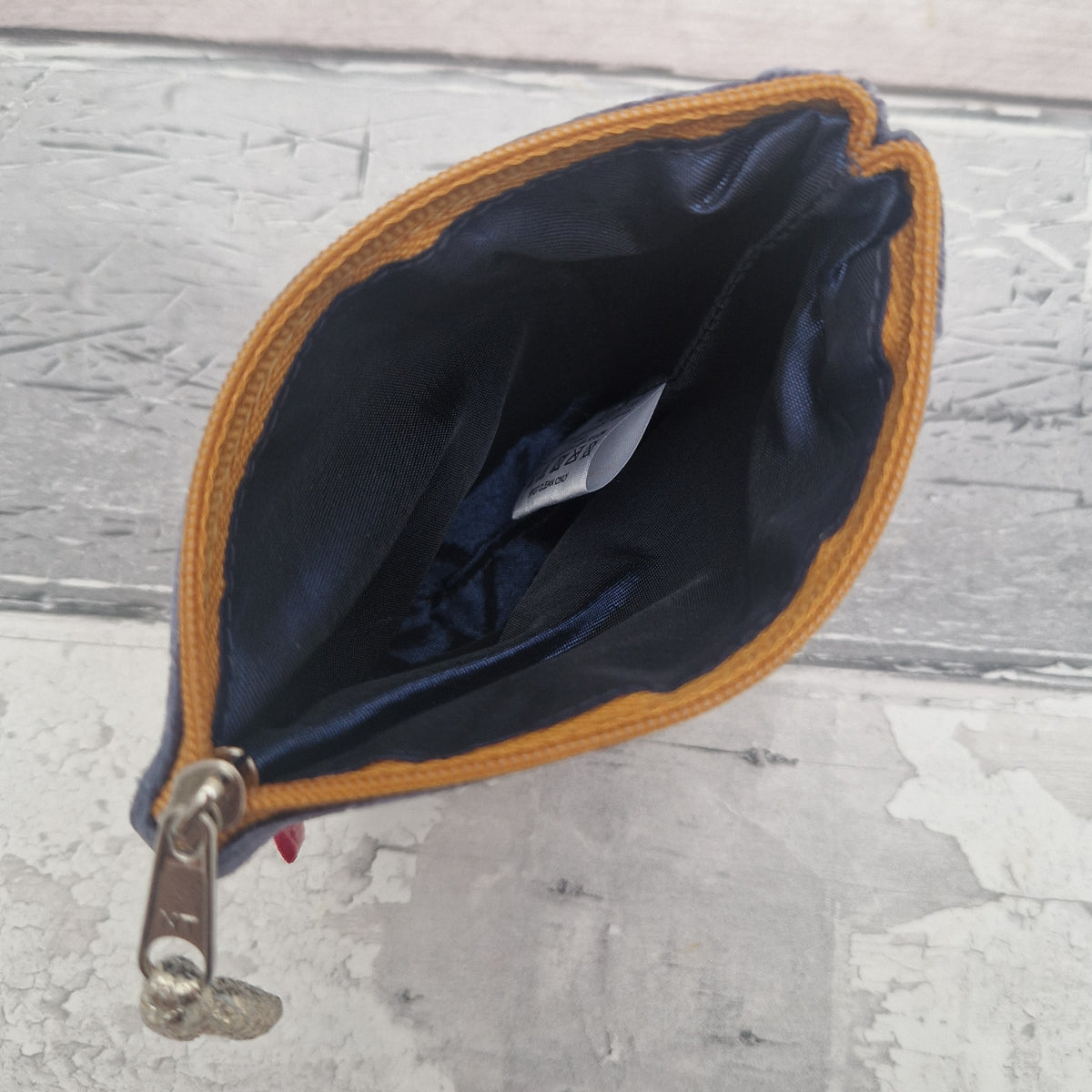 An open zippered pouch or coin purse with a dark navy blue satin-like interior and a mustard yellow fabric trim, photographed from above on a distressed gray surface.