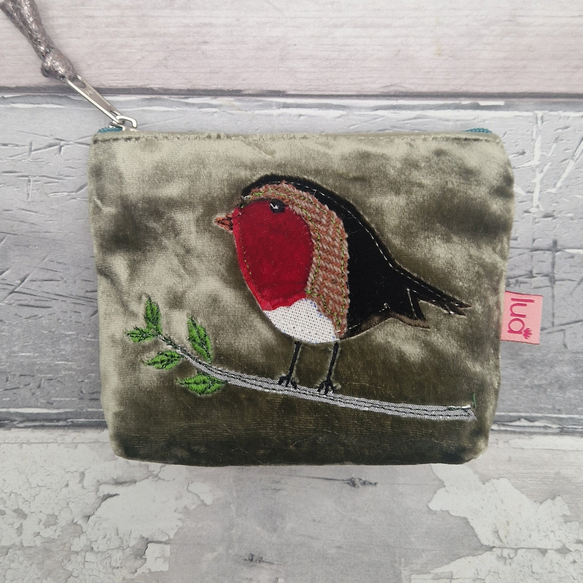 An olive green velvet coin purse featuring an embroidered robin bird on a branch. The coin purse has a silver zipper with a matching strap and a small pink tag on the side.