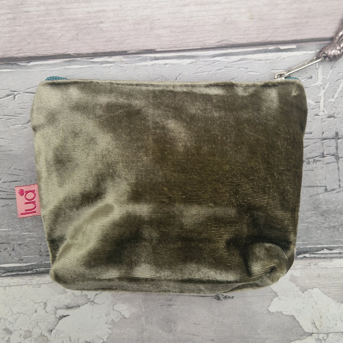 An olive green velvet zippered pouch with a small pink 'oni' brand tag, resting on a distressed gray wooden surface.