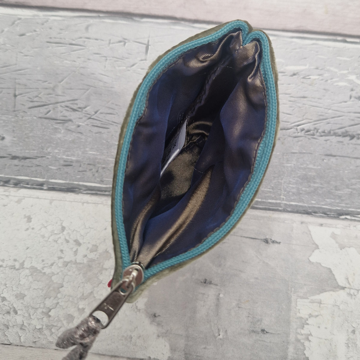 A small zippered pouch with a curved, leaf-like silhouette, featuring a dark navy blue satin-like interior and a teal blue fabric trim along the zipper. The pouch is shown open, revealing its interior, and is photographed on a distressed gray wooden surface.