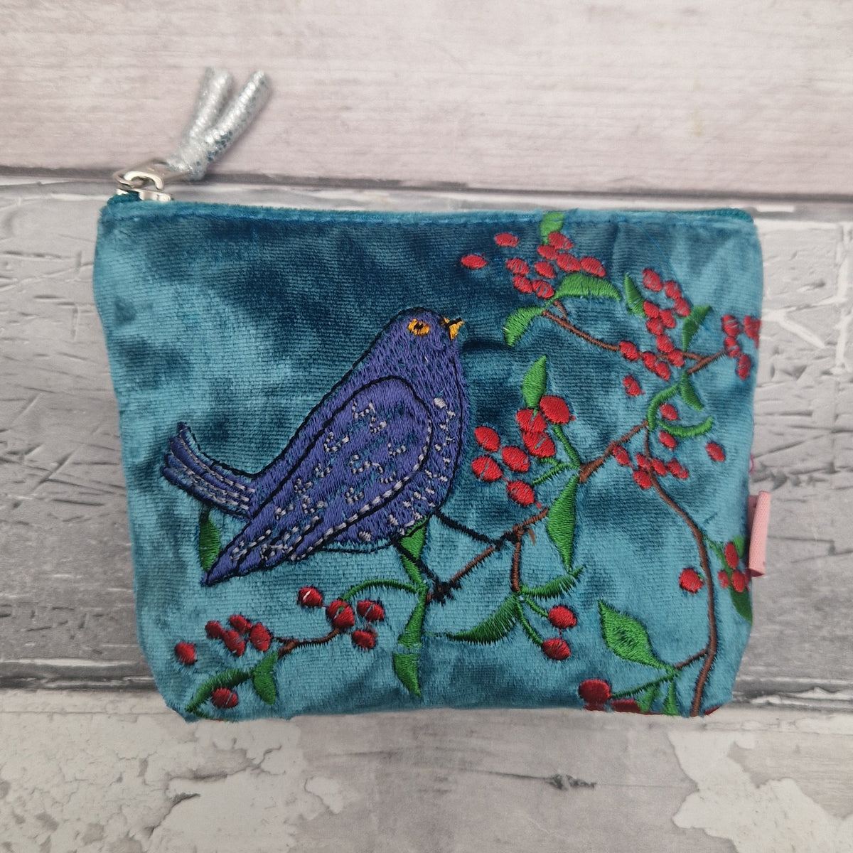 A teal velvet zip pouch featuring an embroidered black bird perched on a branch with red berries, set against a rustic light gray wood background.