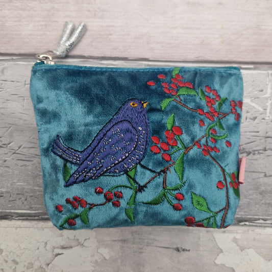 A teal velvet zip pouch featuring an embroidered black bird perched on a branch with red berries, set against a rustic light gray wood background.