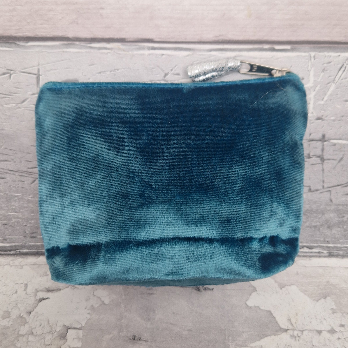 A teal blue velvet zippered pouch with a silver metal zipper pull, resting on a distressed light gray wooden surface.