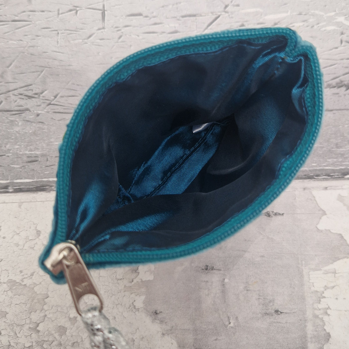A small teal fabric pouch with a satin lining, shown open with a silver zipper pull on a distressed gray background.