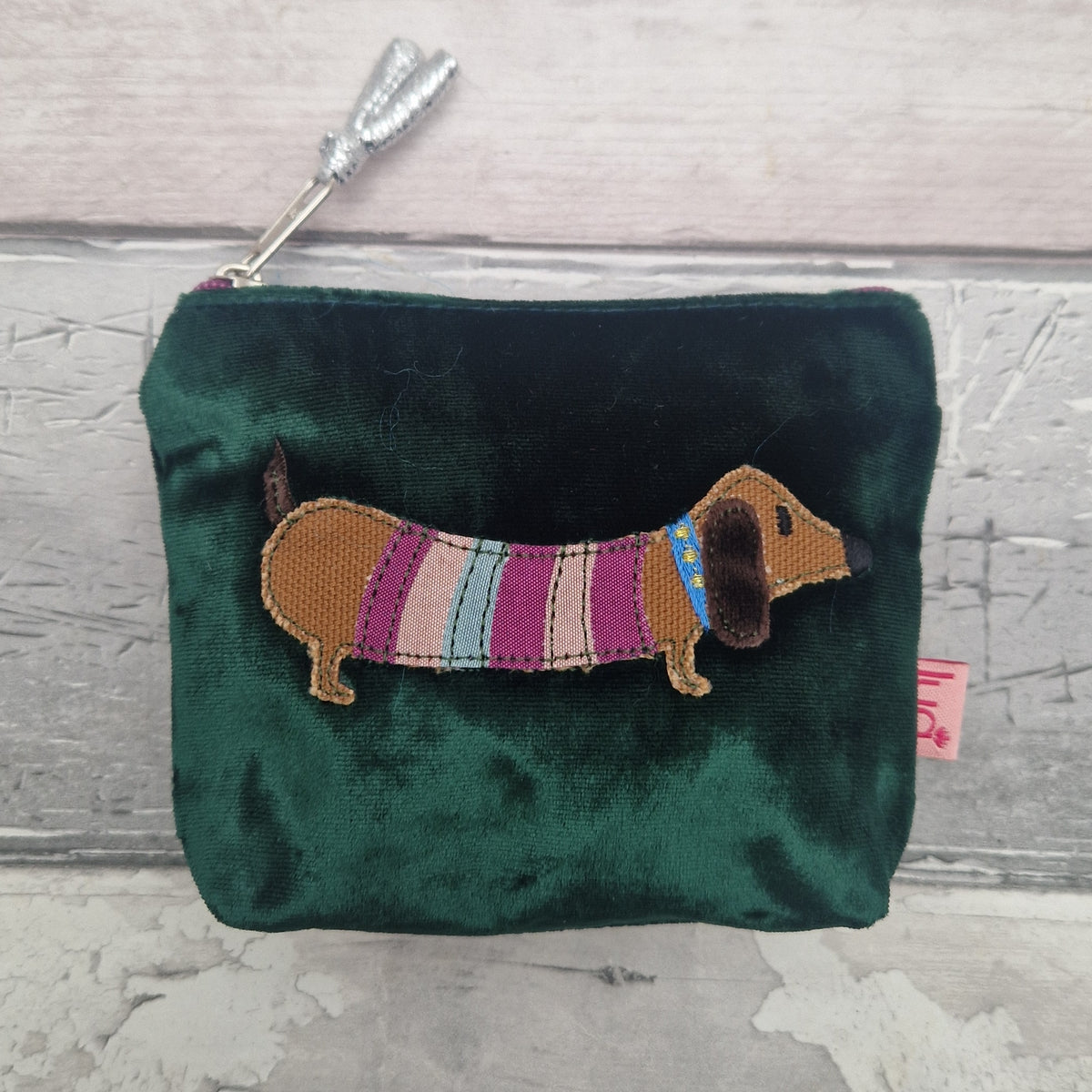 A dark green velvet coin purse featuring an embroidered dachshund wearing a striped sweater, set against a light gray wood plank background.