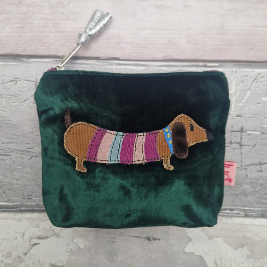 A dark green velvet coin purse featuring an embroidered dachshund wearing a striped sweater, set against a light gray wood plank background.