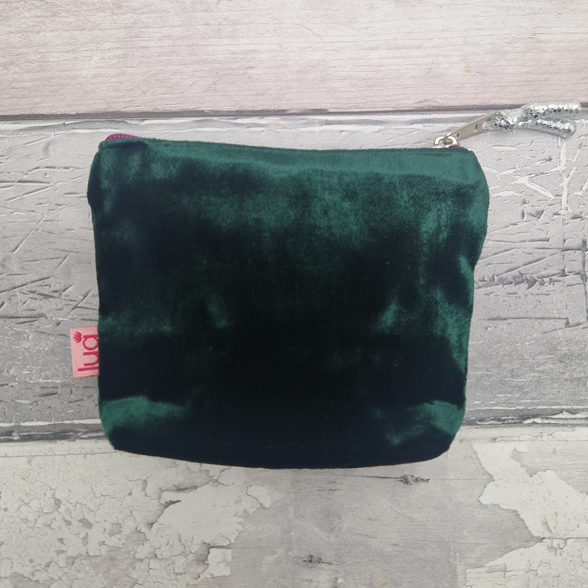 A small dark green velvet zippered pouch with a silver zipper pull, resting on a distressed gray wooden surface. A small pink tag with the brand name 'luA' is visible on the left side.