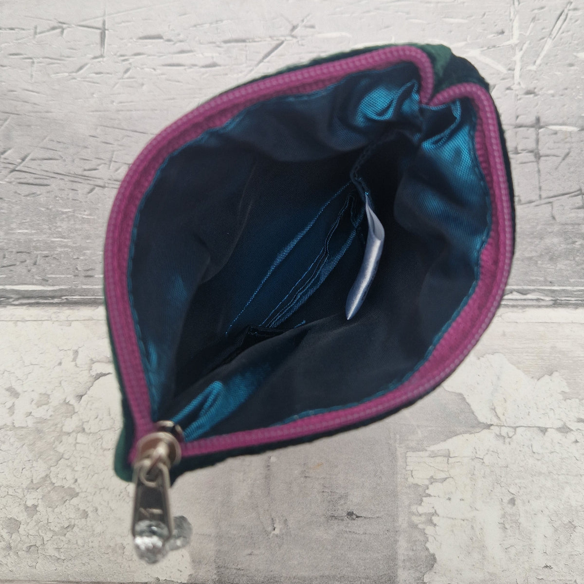 An open zippered pouch with a dark teal satin interior, a magenta-pink stitched edge, and a silver metal snap closure, shown against a textured gray background.