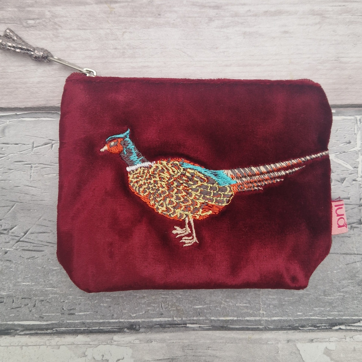 A red velvet zip pouch featuring an embroidered pheasant bird on the front, set against a gray wood-plank background.