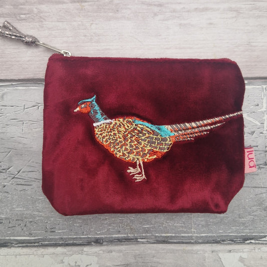 A red velvet zip pouch featuring an embroidered pheasant bird on the front, set against a gray wood-plank background.