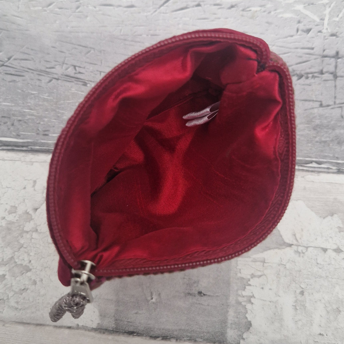 A close-up of a red satin zippered pouch or cosmetic bag, showing the interior lining and a silver zipper pull, set against a textured gray background.
