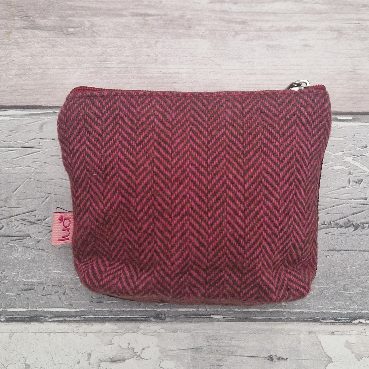 A small zippered pouch in a dark red and pink herringbone tweed fabric, featuring a small pink tag on the left side, set against a rustic gray wooden background.