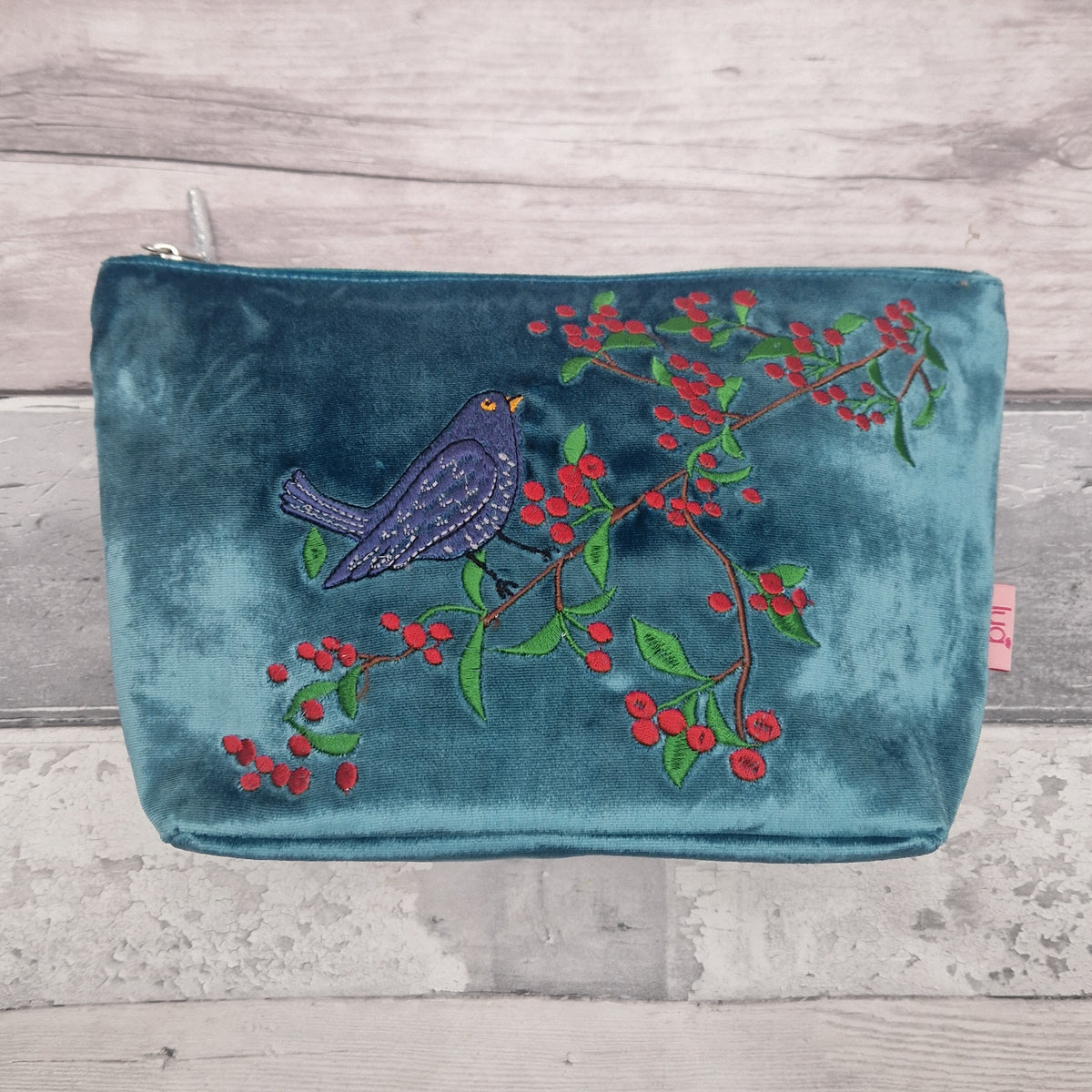 A teal velvet zippered pouch featuring an embroidered design of a blue bird perched on a branch with red berries and green leaves, displayed on a light gray wood plank background.