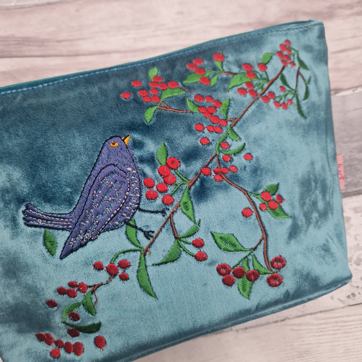 A teal velvet clutch bag featuring an embroidered purple bird perched on a branch with red berries and green leaves, shown on a light wood background.