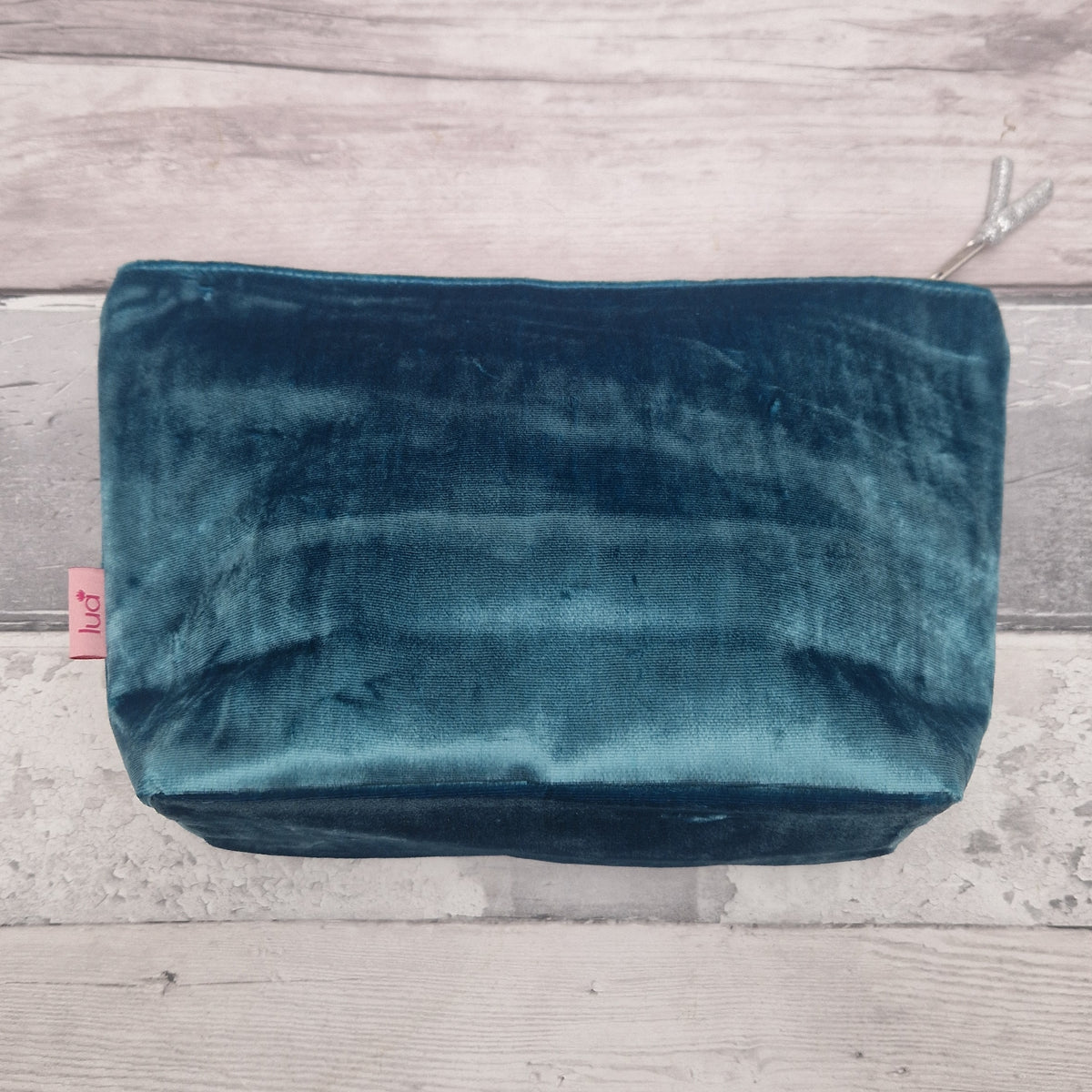 A teal velvet cosmetic pouch with a top zipper, resting on a light gray wooden plank surface. A small pink tag on the left edge displays the brand name 'LUA'.