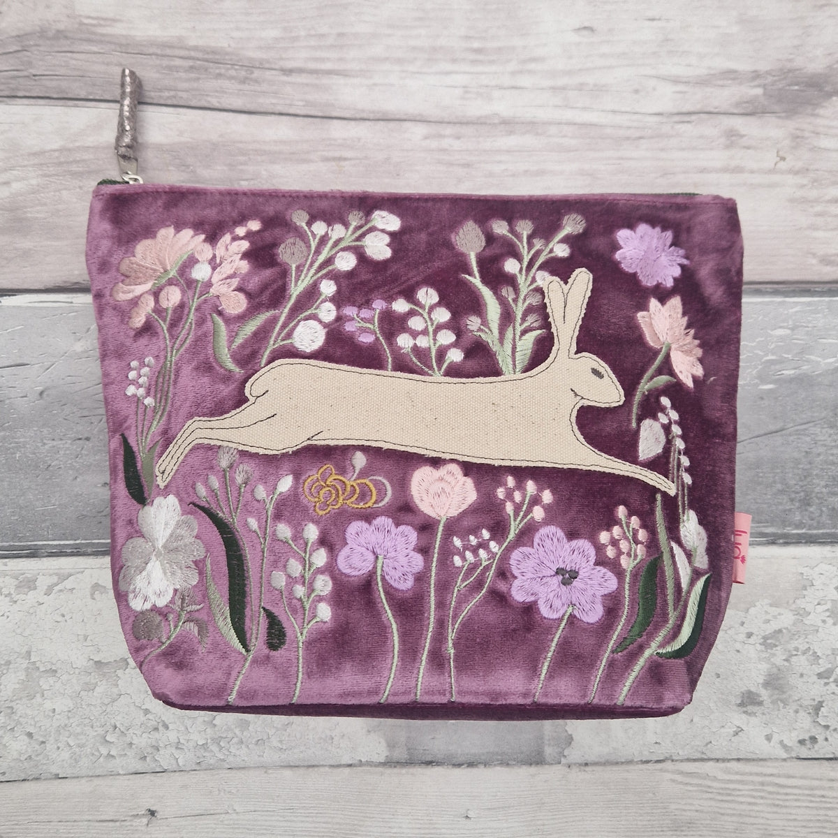 A rectangular purple velvet pouch with a silver zipper pull, decorated with an embroidered design of a beige hare surrounded by pink, purple, and white wildflowers, set against a light gray wood background.