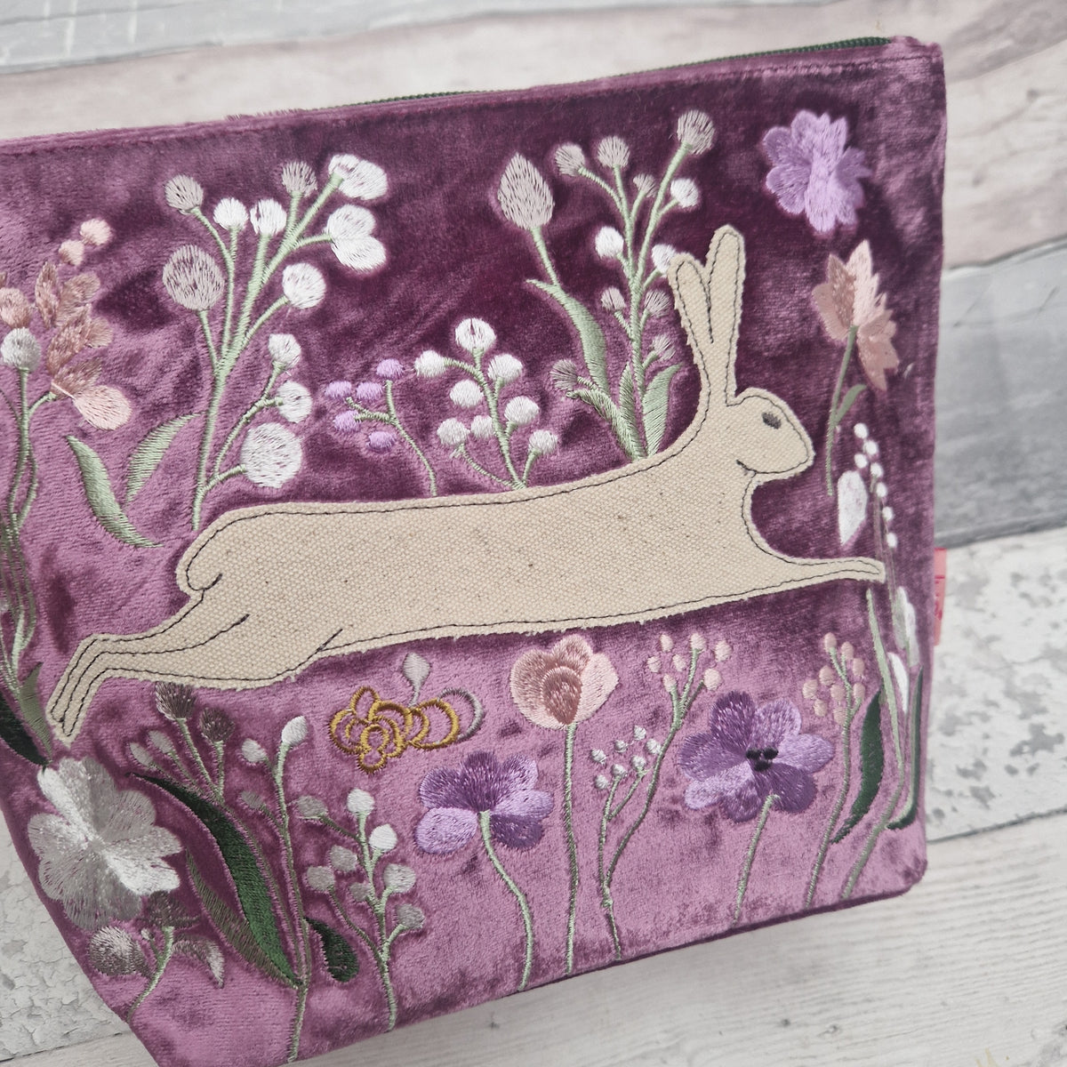 A purple velvet zip pouch featuring an embroidered beige hare and purple, pink, and white wildflowers on a light wood background.