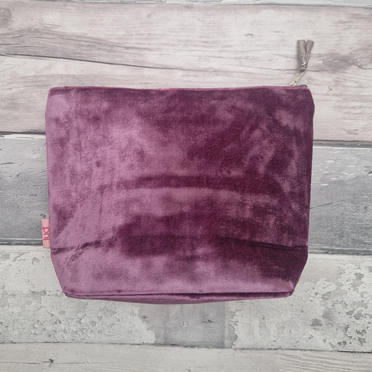 A dusty purple velvet cosmetic pouch with a silver zipper, laid flat on a rustic gray wooden plank background.