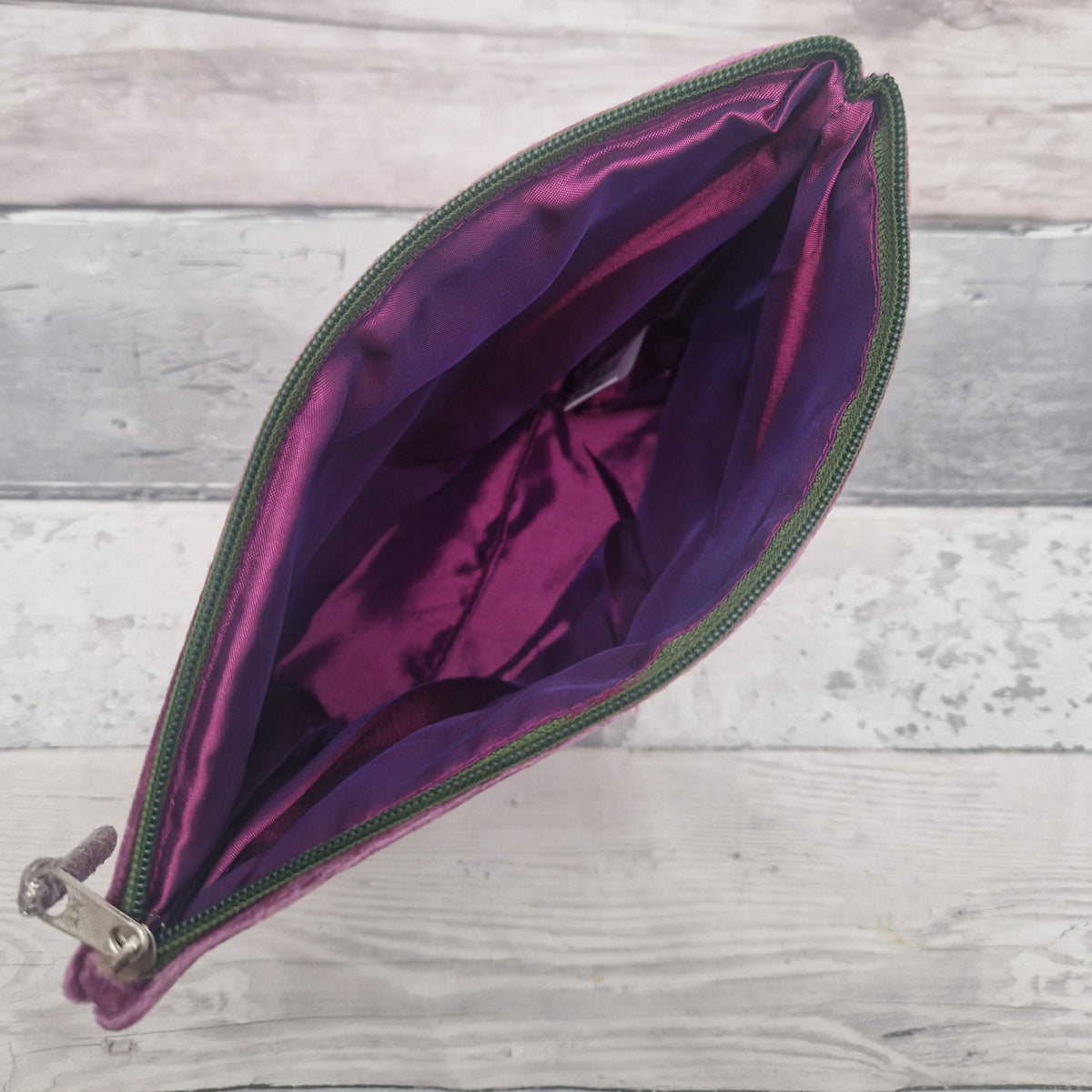 An open zippered pouch with a shiny purple interior and a green zipper, photographed from above on a light gray wood plank background.