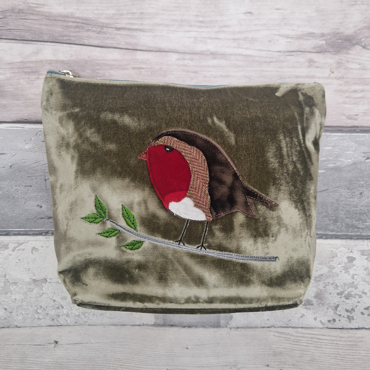 An olive green velvet zippered pouch featuring an embroidered robin bird on a branch, set against a light gray wood plank background.