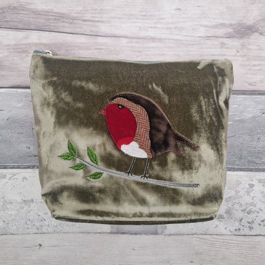 An olive green velvet zippered pouch featuring an embroidered robin bird on a branch, set against a light gray wood plank background.