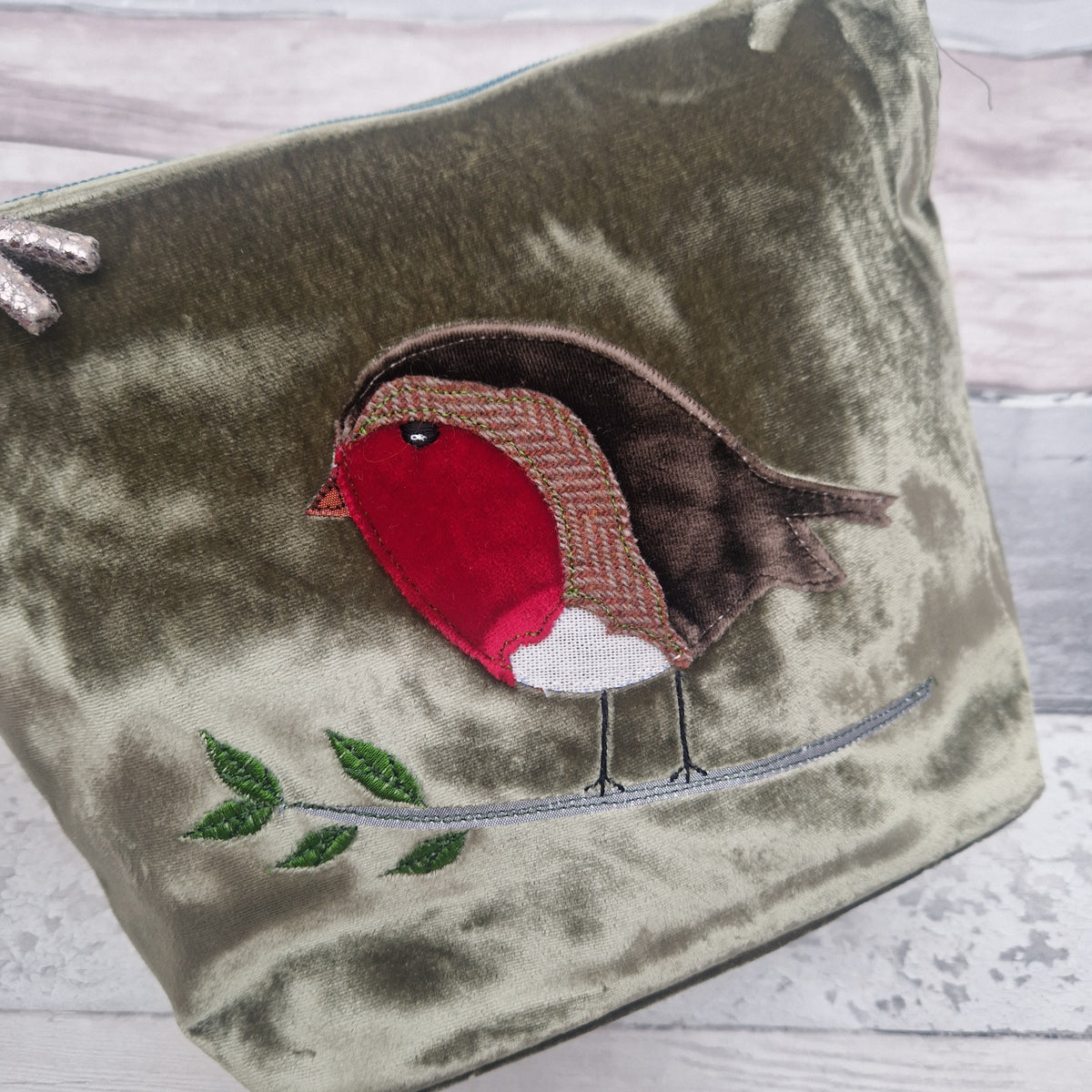 An olive green velvet zippered pouch featuring a detailed robin bird applique on the front, perched on a branch with green embroidered leaves, set against a light gray wooden background.