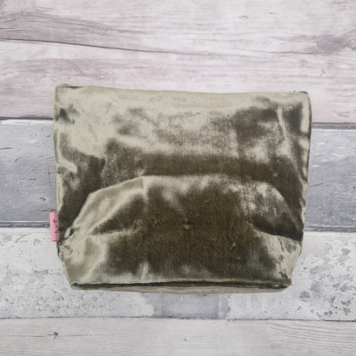 A rectangular olive green velvet pouch or cosmetic bag with a small pink tag on the left side, photographed from above on a light gray wood plank surface.