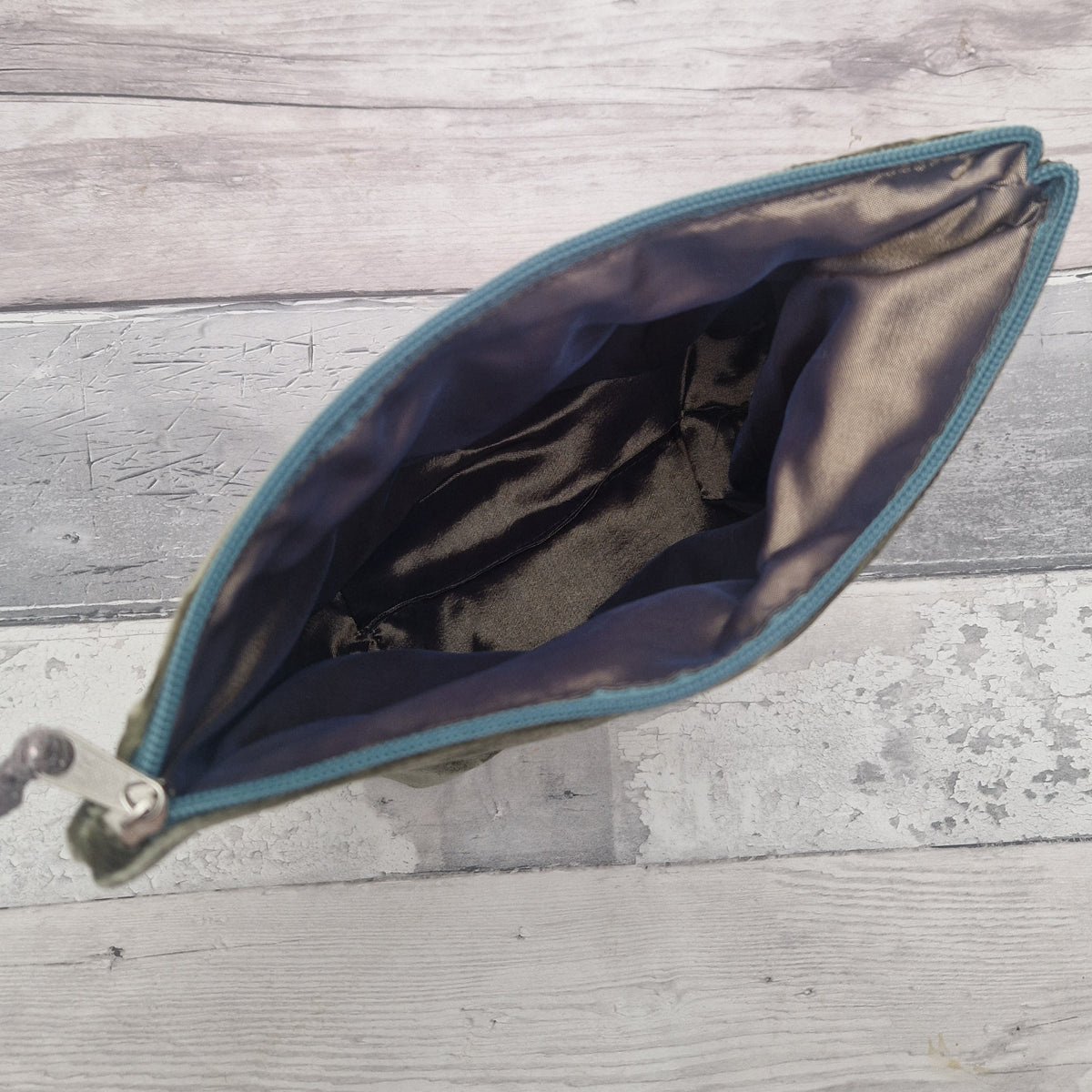 An open zippered pouch or cosmetic bag shown from above on a light gray wood plank surface. The exterior appears olive green or dark khaki, while the interior lining is a shiny dark brown or black satin-like material with a teal zipper edge.