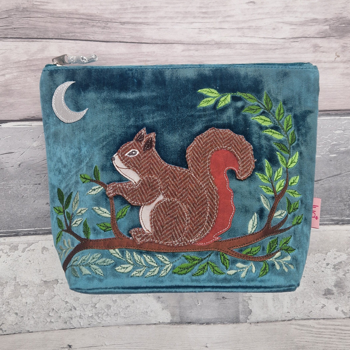 A teal velvet zippered pouch featuring an embroidered brown and red squirrel on a tree branch with green leaves, set against a rustic gray wood plank background.