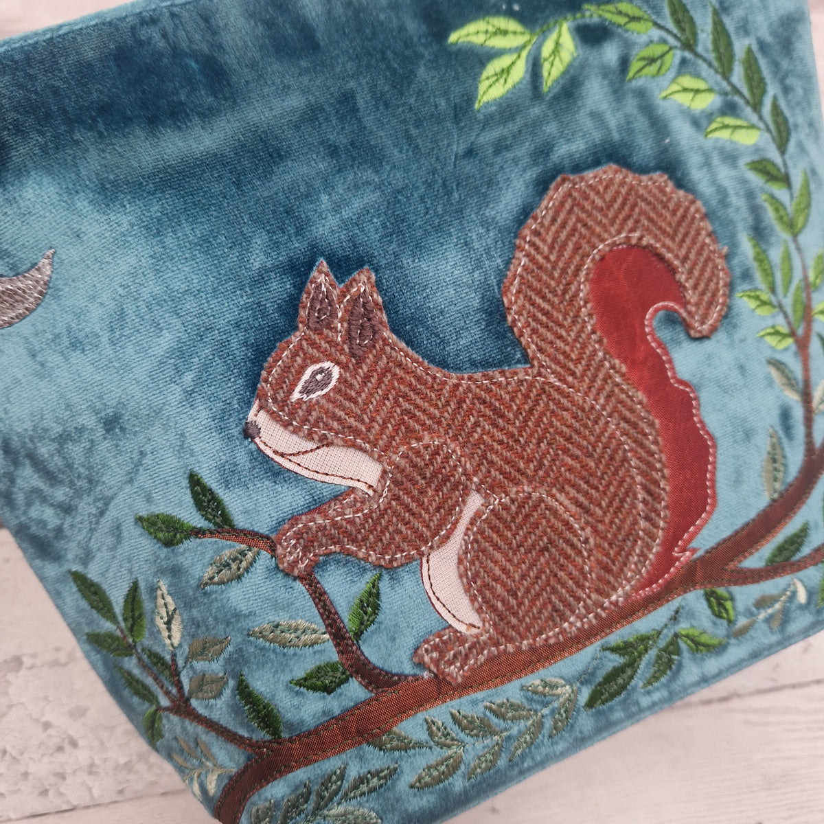 A close-up of a teal velvet fabric item featuring an embroidered brown and red squirrel on a tree branch with green leaves.