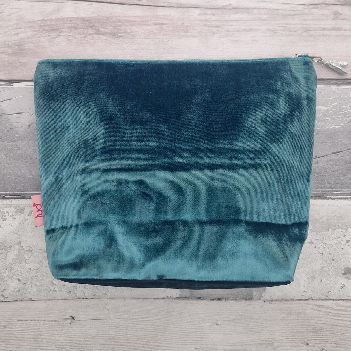 A teal velvet cosmetic pouch with a silver zipper, laid flat on a light gray wood plank background.
