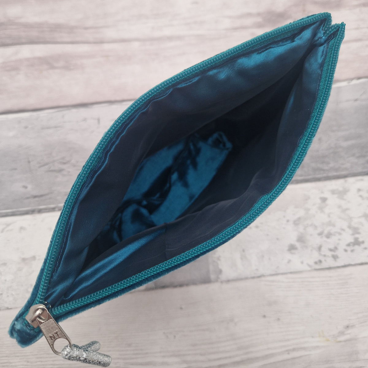 An open teal zippered pouch with a shiny teal satin lining, shown from a top-down view on a light gray wood plank background.