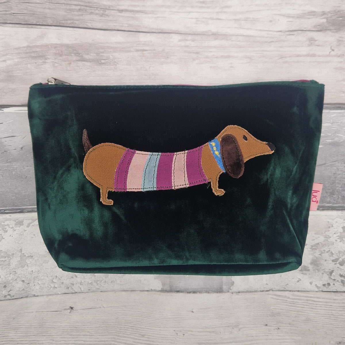 A dark green velvet zippered pouch featuring an embroidered dachshund wearing a pink, purple, and blue striped sweater, set against a light gray wood plank background.